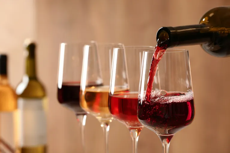Rotwein wird in eines von mehreren Gläsern eingeschenkt, im Hintergrund sind weitere Rot- und Weißweinflaschen auf einer unscharfen Holzfläche zu sehen.