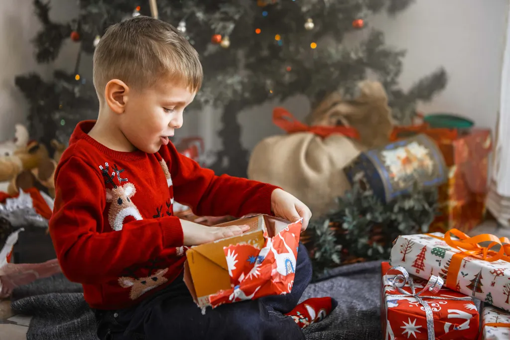 Kleiner Junge im roten Weihnachtspullover sitzt vor einem geschmückten Christbaum und packt neugierig ein Weihnachtsgeschenk aus. Neben ihm liegen weitere verpackte Geschenke