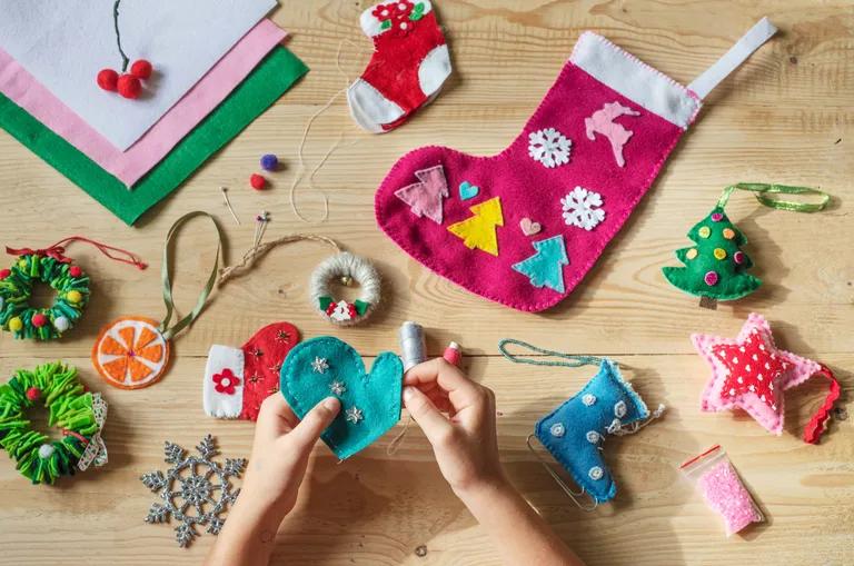 Hände, die auf einem Holztisch bunte Weihnachtsschmuckstücke aus Filz basteln, darunter Strümpfe, Sterne und Tannenbäume.