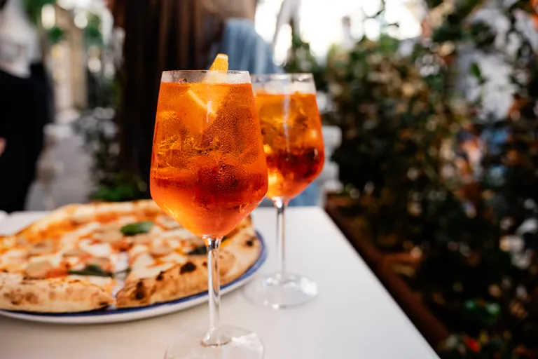 Zwei Gläser Aperol Spritz mit Orangenscheiben auf einem Tisch, dazu eine frisch gebackene Pizza im Freien.