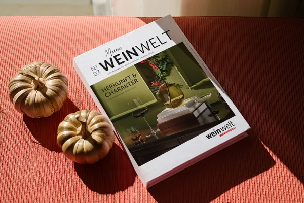 Ein Tisch mit orangefarbener Tischdecke, auf der zwei Dekokürbisse links neben einer Ausgabe des "Meine weinwelt"-Magazins Herbst 2026 liegen.