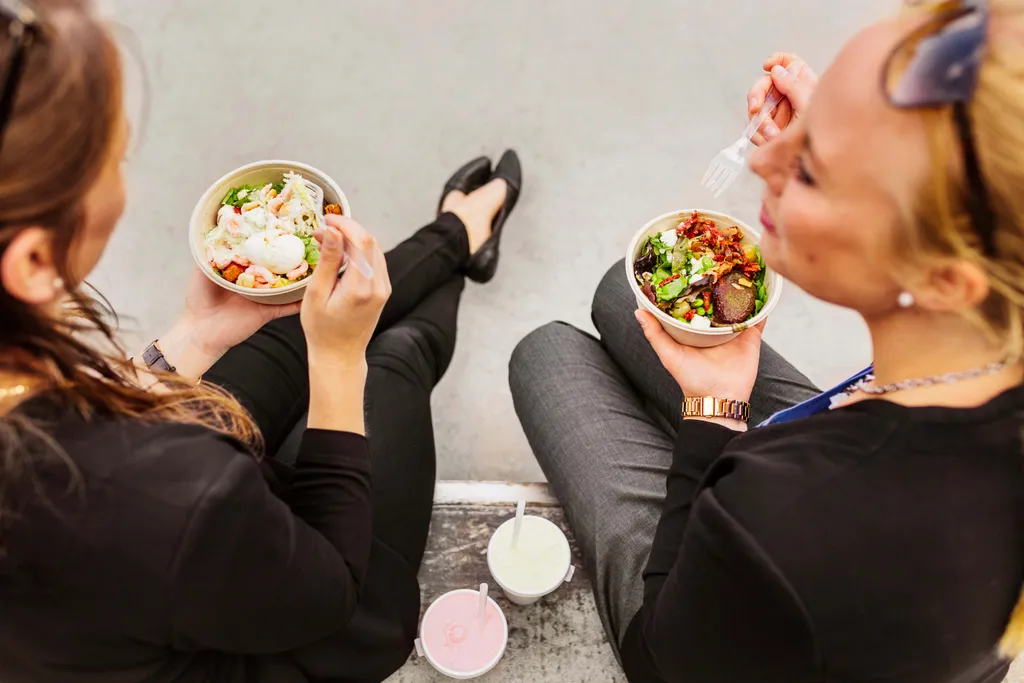Zwei Frauen sitzen im Freien und genießen ihre Salate, während ihre Getränke neben ihnen auf dem Boden stehen.