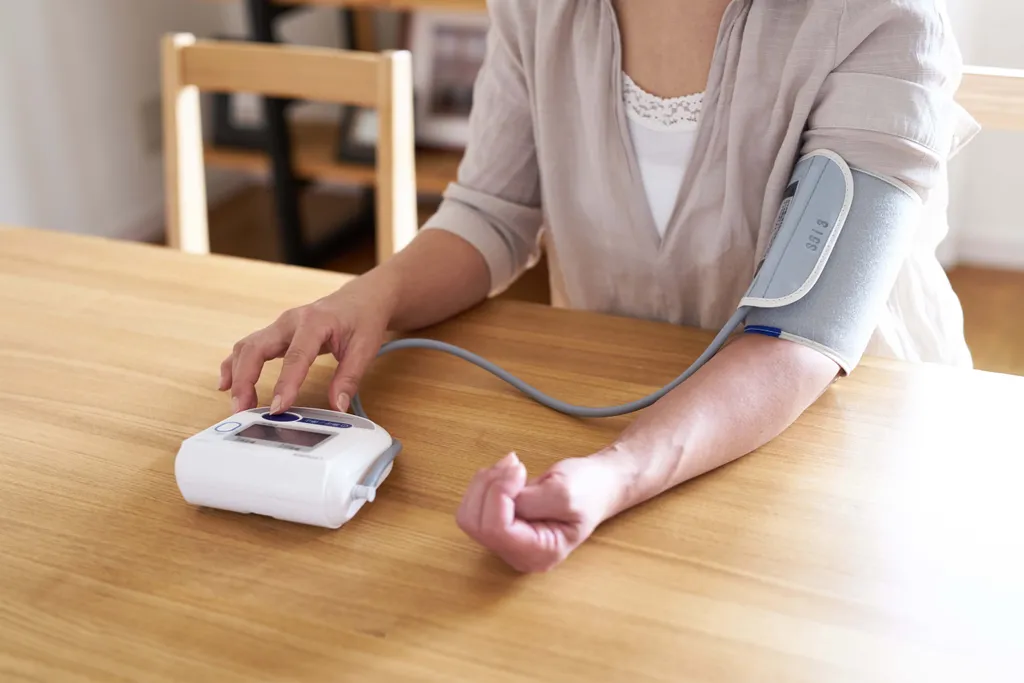 Person misst ihren Blutdruck mit einem digitalen Oberarm-Blutdruckmessgerät am Esstisch zu Hause