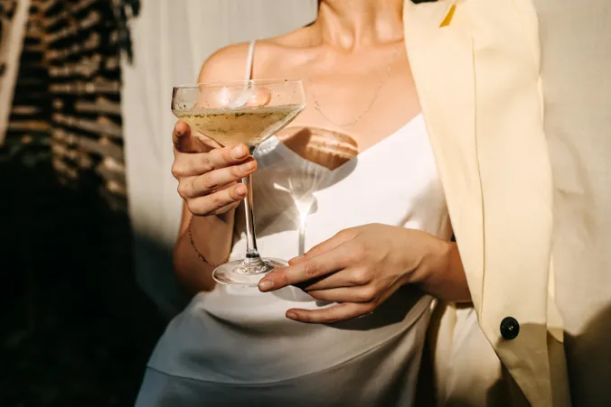 Eine Person in einem weißen Kleid und einem beigen Blazer hält ein Cocktailglas in der Hand, während das Sonnenlicht Schatten wirft.