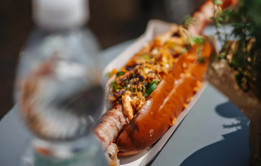 Ein reichlich belegter Hotdog liegt in einer Papierschale auf einem Tisch neben einer verschwommenen Wasserflasche und einer Pflanze.