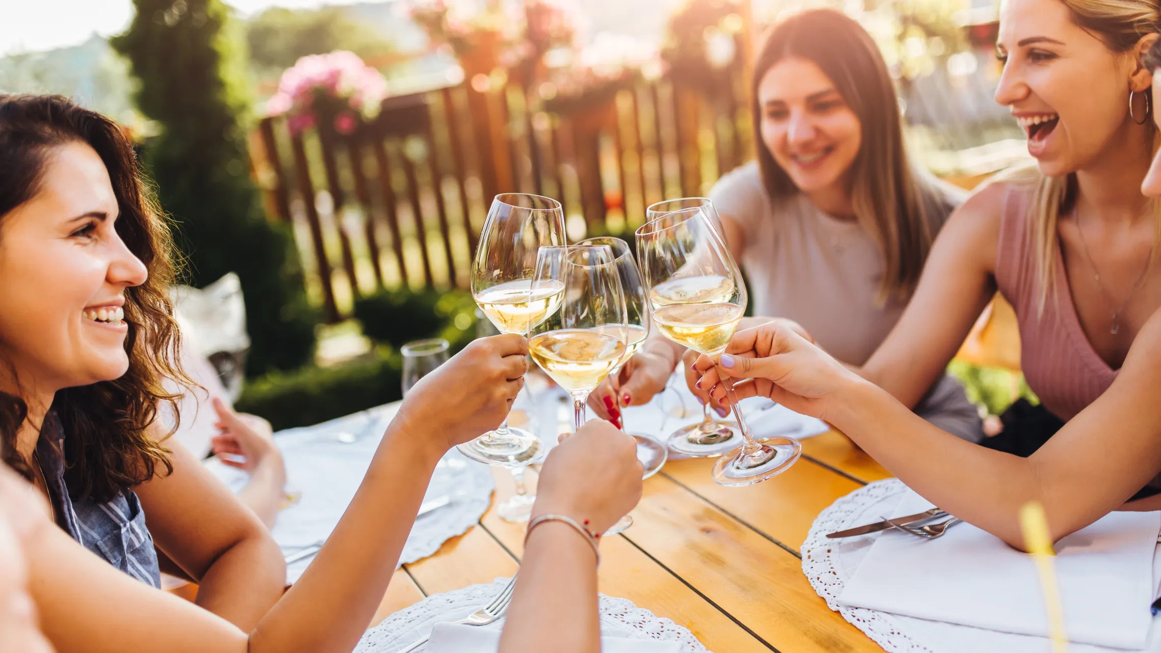 Frauen, die an einem Tisch im Freien mit Weingläsern anstoßen, lachen und die gemeinsame Zeit in einem sonnigen Garten genießen.