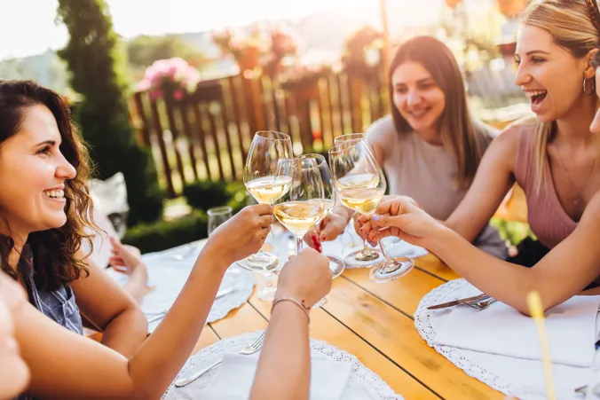 Frauen, die an einem Tisch im Freien mit Weingläsern anstoßen, lachen und die gemeinsame Zeit in einem sonnigen Garten genießen.