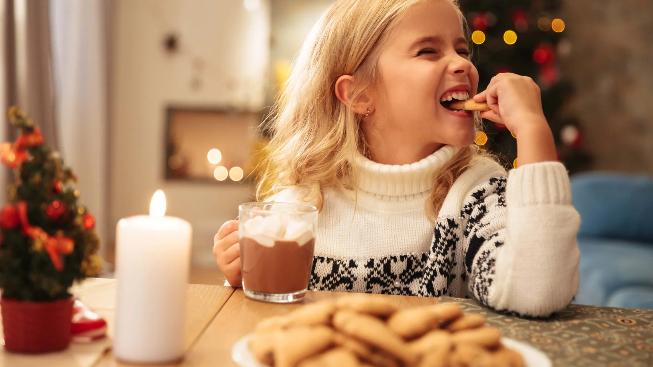 Ein lachendes Mädchen genießt einen Keks und hält eine Tasse heißen Kakao mit Marshmallows. Vor ihr steht ein Teller mit Keksen.