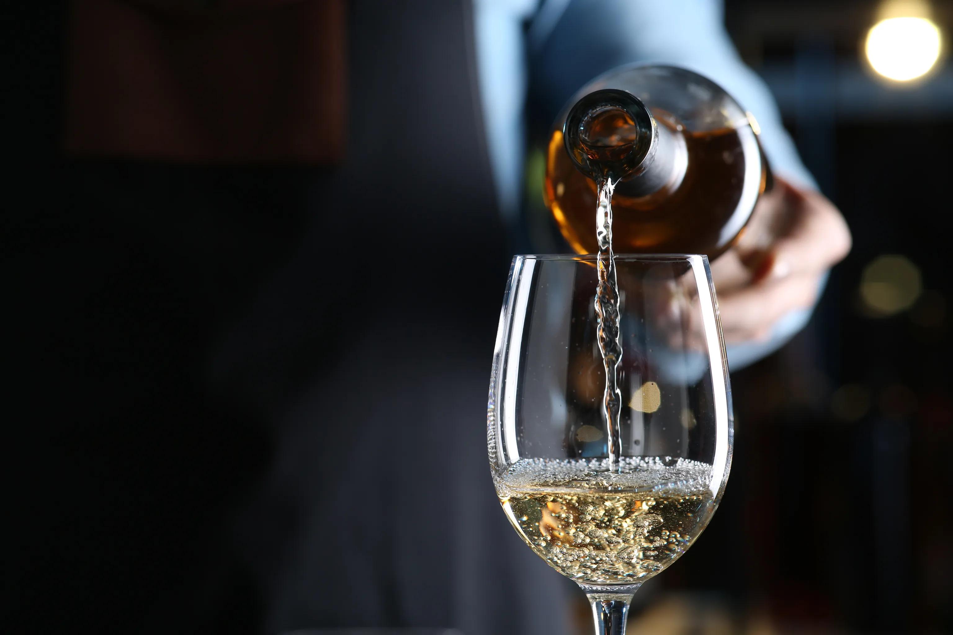 Vor einem verschwommenen dunklen Hintergrund gießt eine Person Weißwein aus einer Flasche in ein Glas, wobei sich an der Oberfläche Bläschen bilden.