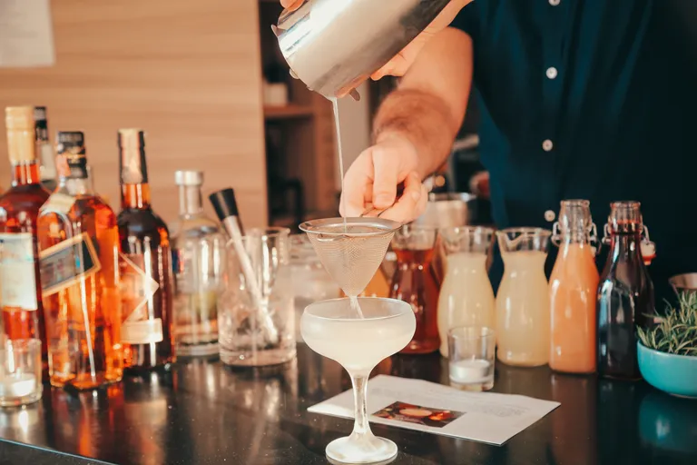 Ein Barkeeper, der einen Cocktail in ein Glas abseiht, umgeben von verschiedenen Spirituosenflaschen und Mixzutaten auf einer Bartheke.