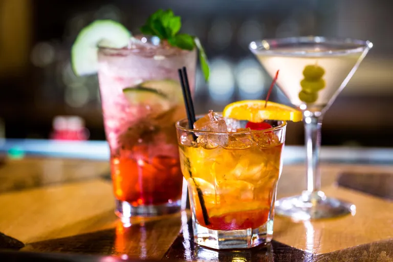 Nahaufnahme von drei Cocktails: ein Whisky mit Orange und Kirsche, ein Martini mit Oliven und ein mit Minze garnierter Mixgetränk auf einem Holztisch.
