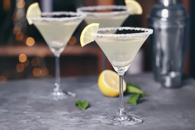 Drei Martini-Gläser mit gesalzenem Rand, gefüllt mit Cocktails, die mit Zitronenschnitzen garniert sind, auf einem Tisch mit einer Zitronenhälfte und Minzblättern.