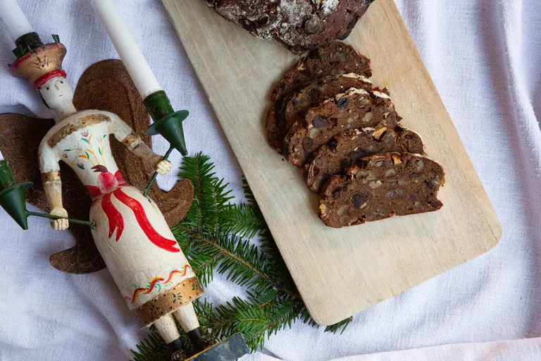 Aufgeschnittenes Kletzenbrot mit Nüssen und Trockenfrüchten auf einem Holzbrett neben Tannenzweigen und einer Weihnachtsfigur