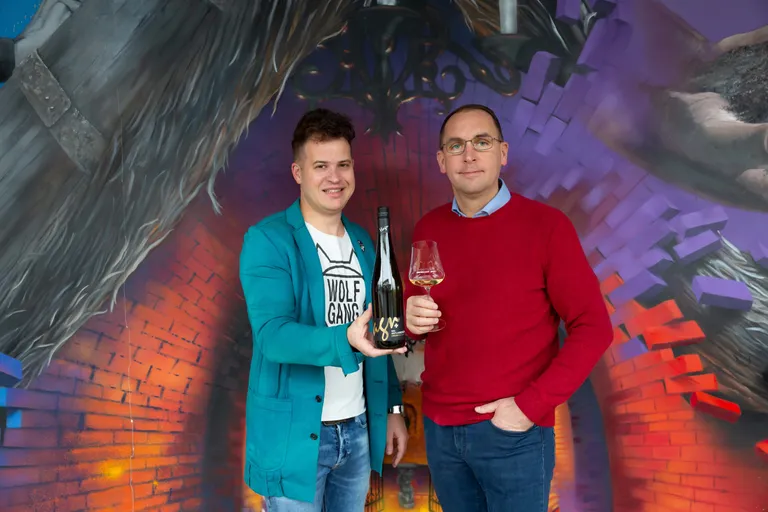 Wolfgang Hagn Jun. und Leo Hagn Jun. vor einer bunten Wand mit einem Weinglas bzw. einer Flasche Wein in den Händen