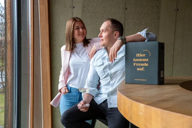 Ein Mann und eine Frau lächeln am Fenster, der Mann hält ein Weinglas in der Hand, auf der Theke steht eine Schachtel mit der Aufschrift „Hier kommt Freude auf“.
