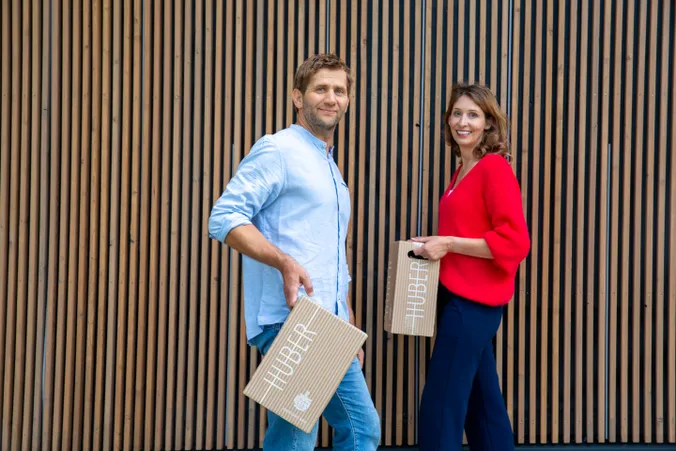 Ein Mann und eine Frau, die in die Kamera lächeln, HUBER-Kartonverpackungen in den Händen halten und vor einer Wand aus Holzpaneelen stehen.