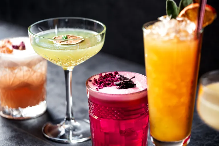 Eine Auswahl an farbenfrohen Cocktails: ein grüner im Coupé-Glas, ein rosa mit Schaum, ein orangefarbener mit Eis und zwei cremige Getränke mit Garnitur.