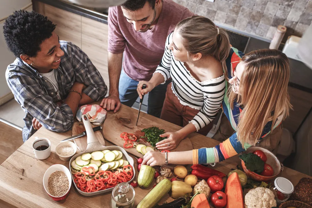 Menschen, die in einer Küche eine Mahlzeit zubereiten; auf einem Holztisch liegen verschiedene Gemüsesorten wie Zucchini, Paprika und Blattgemüse sowie Tassen und Schüsseln.