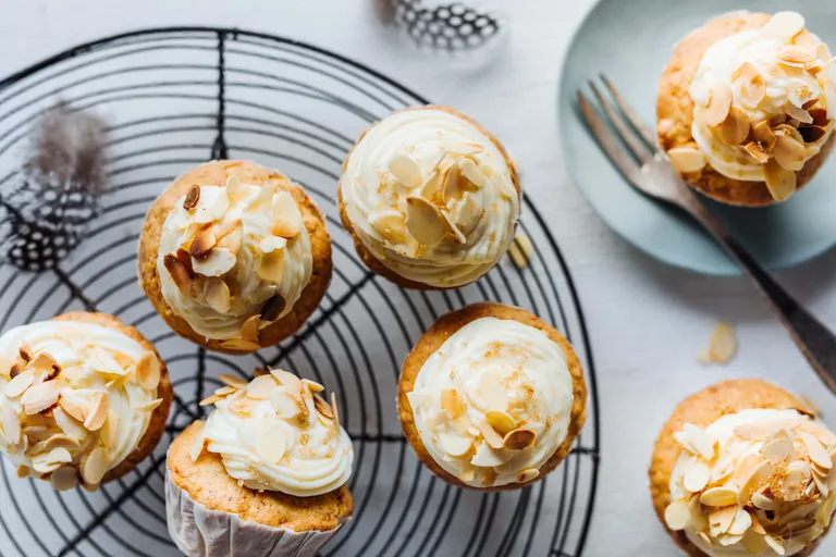 Cupcakes mit Sahne und Mandelscheiben auf einem Kuchengitter, daneben eine Gabel auf einem kleinen blauen Teller.