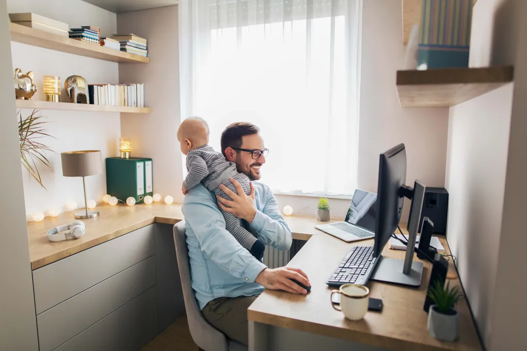 Vater arbeitet im Homeoffice und hält gleichzeitig sein Baby auf dem Arm