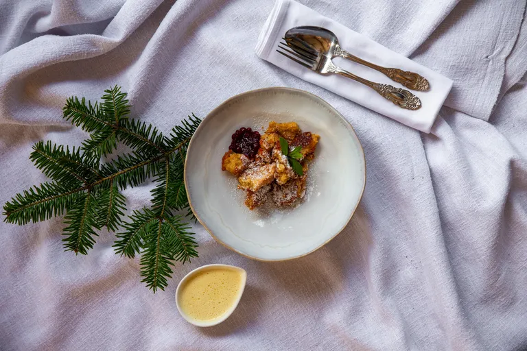Lebkuchen-Kaiserschmarrn mit Staubzucker und Preiselbeeren auf einem Teller, dazu Vanillesauce und Tannenzweige als winterliche Dekoration