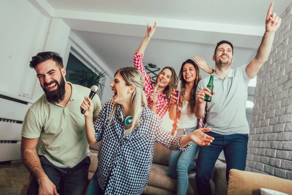 5 glückliche Freunde haben Spaß, singen Karaoke und fühlen die Musik. Ein Teil sitzt auf der Couch und hat Flaschen in der Hand, der andere Tei kniet auf dem Boden im Wohnzimmer.