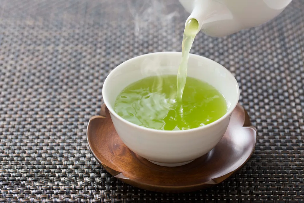 Leuchtend grüner Tee wird in eine weiße Tasse auf einer hölzernen Untertasse gegossen, die auf einer geflochtenen Unterlage steht.