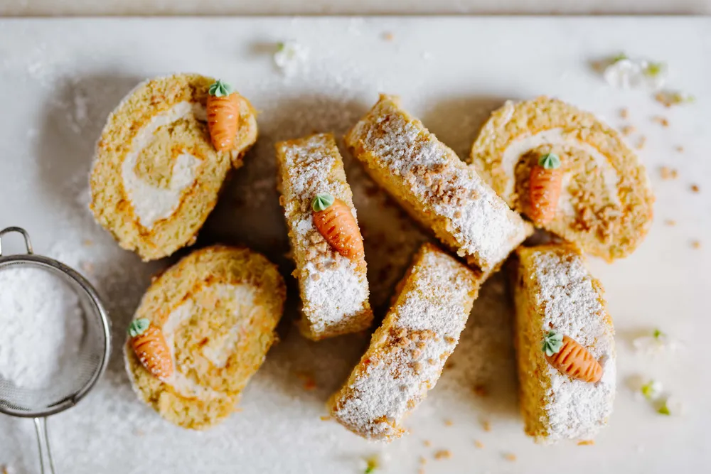 Eine in Scheiben geschnittene Karottenkuchenrolle mit Cremefüllung, garniert mit kleinen Karottenfiguren und mit Puderzucker bestäubt, präsentiert auf einer weißen Unterlage.
