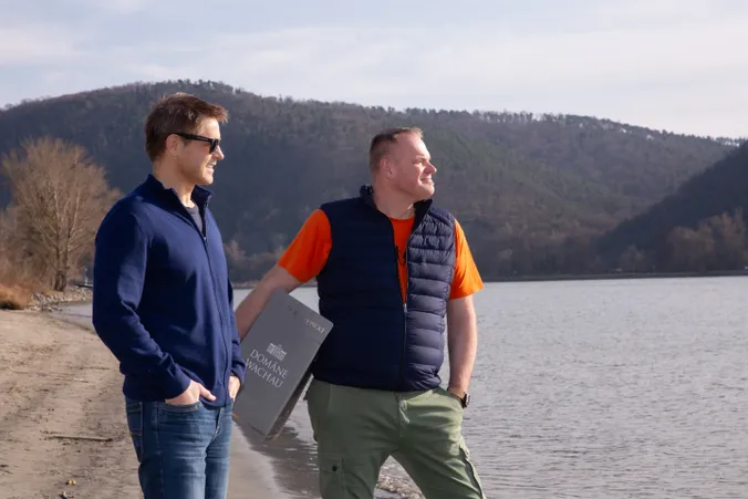Zwei Männer stehen am Seeufer, einer von ihnen hält eine Weinkiste der Marke „Domäne Wachau“ in der Hand. Im Hintergrund ist unter einem teilweise bewölkten Himmel ein bewaldeter Hügel zu sehen.