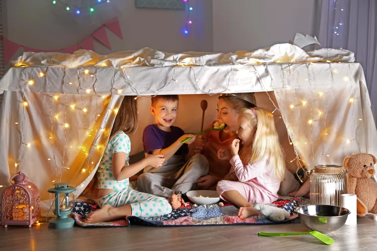 Drei Kinder und eine Mutter sitzen in einer selbst gebauten Höhle aus Decken und Kissen im Wohnzimmer