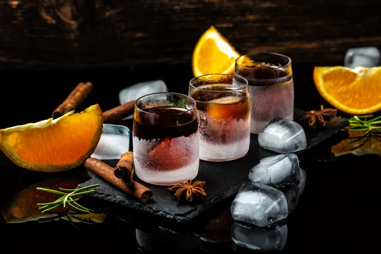 Beschlagene Gläser mit dunklem Alkohol auf einer Schieferplatte, umgeben von Eiswürfeln, Orangenscheiben, Zimtstangen und Rosmarinzweigen.