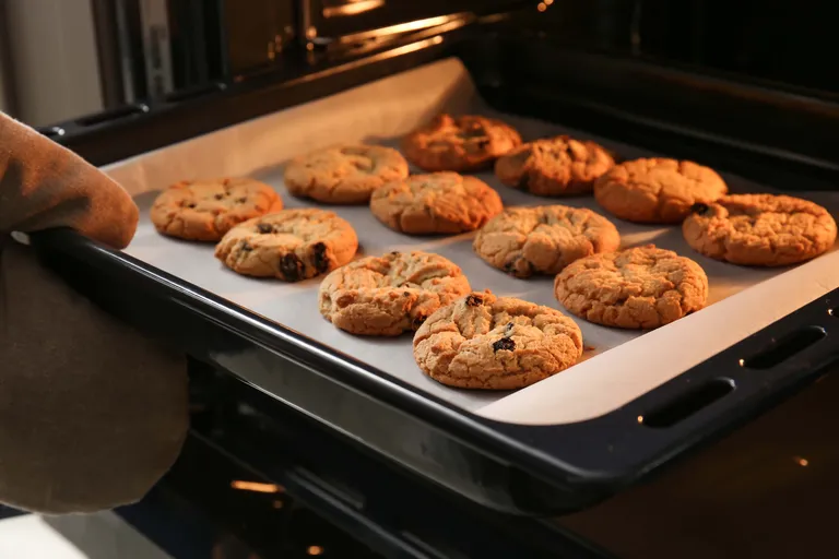 Ein mit Backpapier belegtes Backblech mit goldbraunen Keksen wird mithilfe eines Ofenhandschuhs aus dem Backofen gezogen.