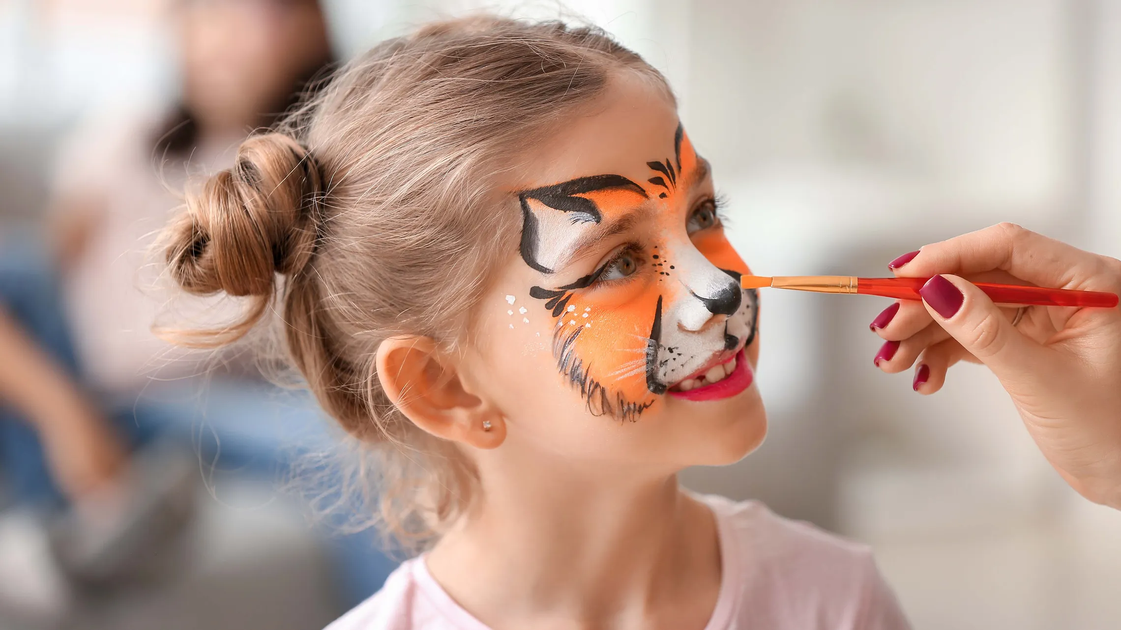 Child with hair in a bun getting a tiger face paint, smiling as an artist applies orange and black paint with a brush.