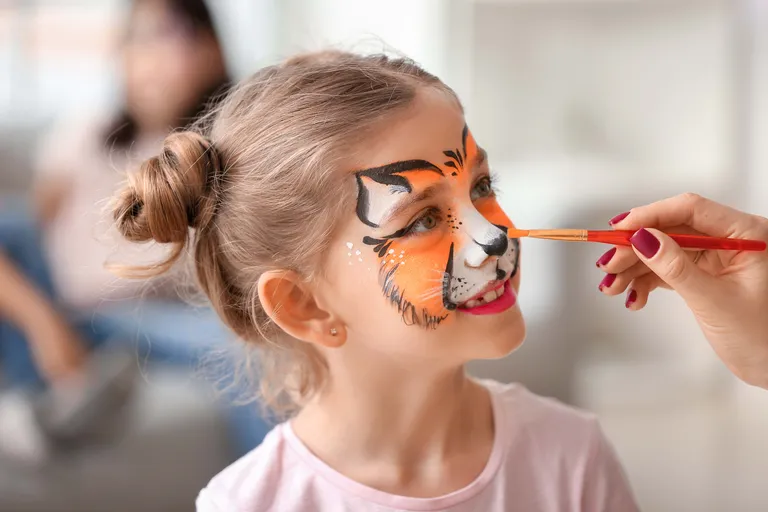 Child with hair in a bun getting a tiger face paint, smiling as an artist applies orange and black paint with a brush.