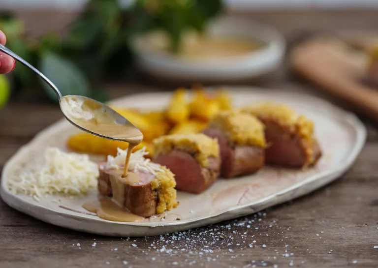 Löffel, der Soße über kleine Schweinefilets auf einem Keramikteller mit geriebenem Kren und Bratkartoffeln gießt.