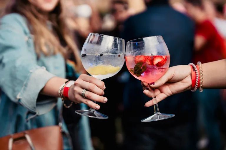 Zwei Menschen stoßen bei einer Veranstaltung im Freien mit Gläsern an, die mit bunten Cocktails gefüllt sind – das eine mit Erdbeeren und Minze, das andere mit Zitrusfrüchten und Beeren.