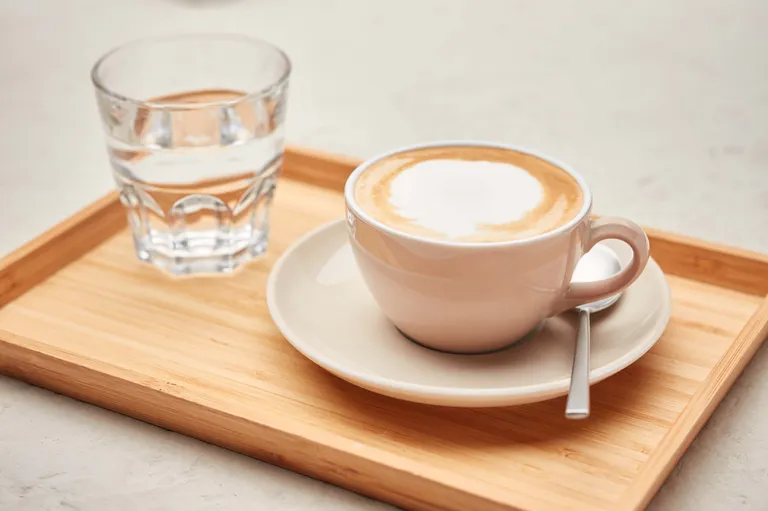 Ein Cappuccino und ein Glas Wasser auf einem Holztablett, im unscharfen Hintergrund eine kleine Topfpflanze.