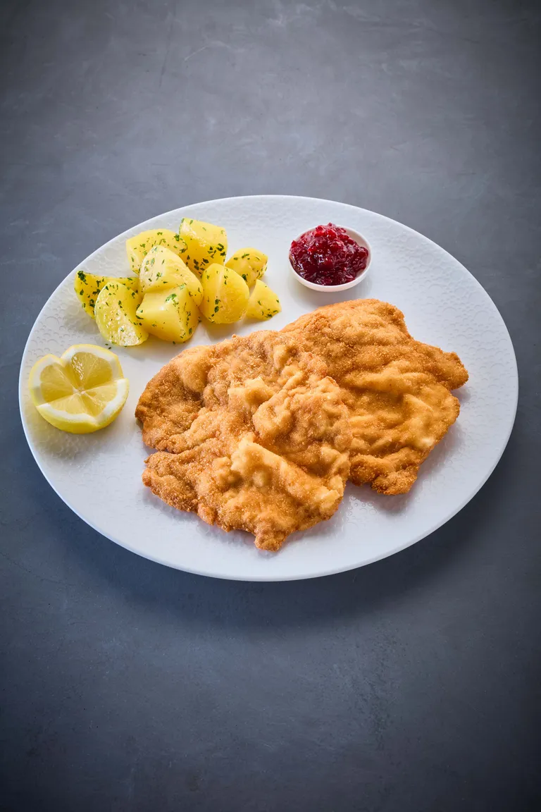 Ein Teller mit Schnitzel, Petersilienkartoffeln, Zitronenscheiben und einer Beilage aus Preiselbeersauce auf einem strukturierten weißen Teller.