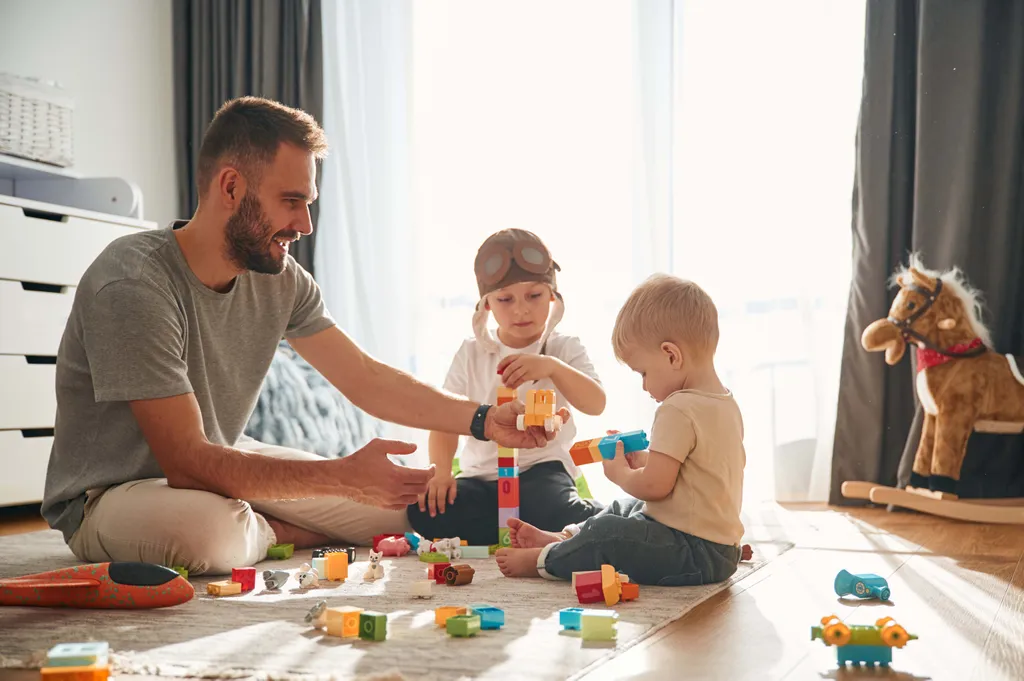 Ein Mann und zwei kleine Kinder spielen auf dem Boden mit bunten Bauklötzen, im Hintergrund steht ein Schaukelpferd.