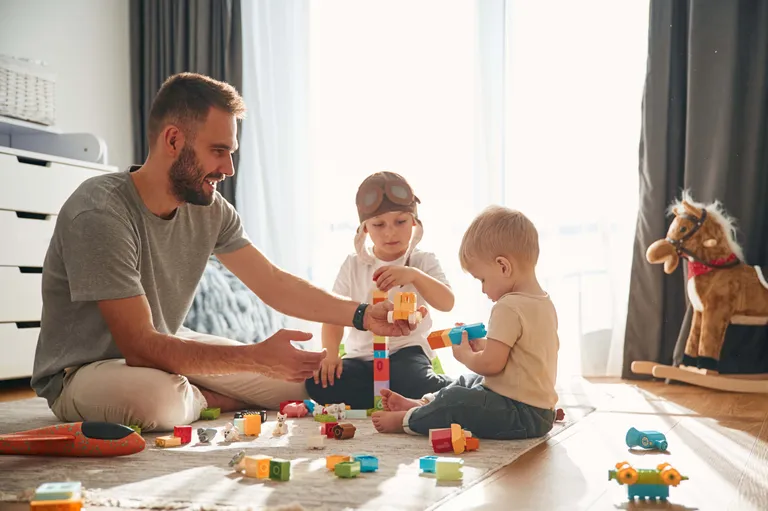 Ein Mann und zwei kleine Kinder spielen auf dem Boden mit bunten Bauklötzen, im Hintergrund steht ein Schaukelpferd.