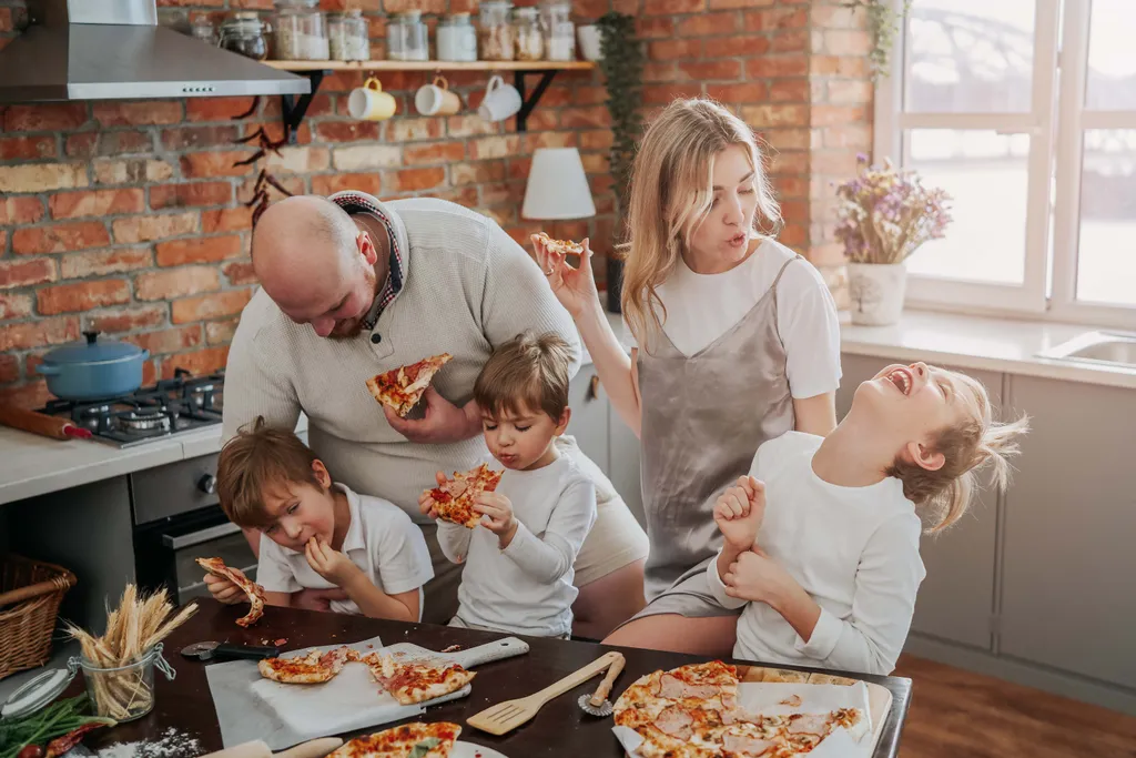 Eine Familie genießt gemeinsam Pizza in einer gemütlichen Küche; voller Lachen und Freude sitzen sie mit einem Stück Pizza in der Hand um den Tisch herum.