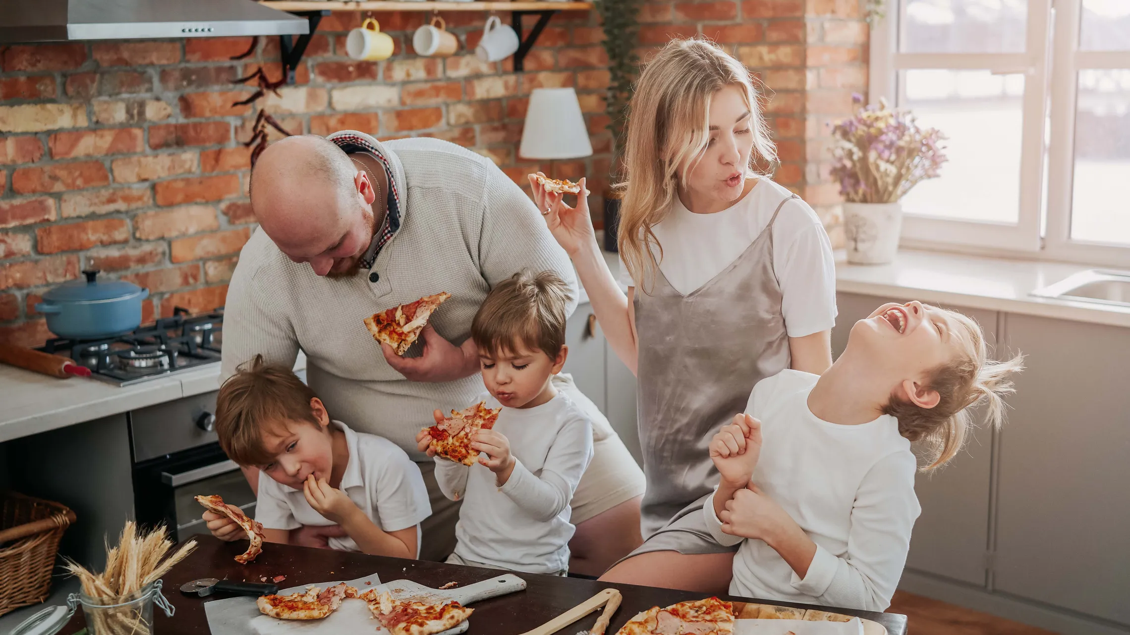 Eine Familie genießt gemeinsam Pizza in einer gemütlichen Küche; voller Lachen und Freude sitzen sie mit einem Stück Pizza in der Hand um den Tisch herum.