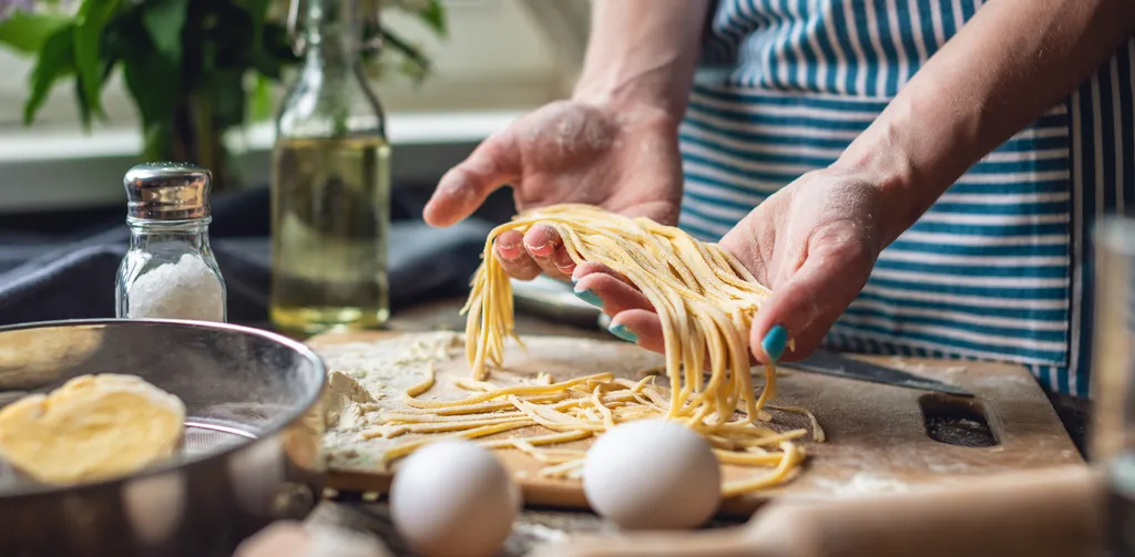 Eine Person macht Pasta selbst, hält Nudeln in den Händen in einer Küche mit Schneidebrettern und Zutaten