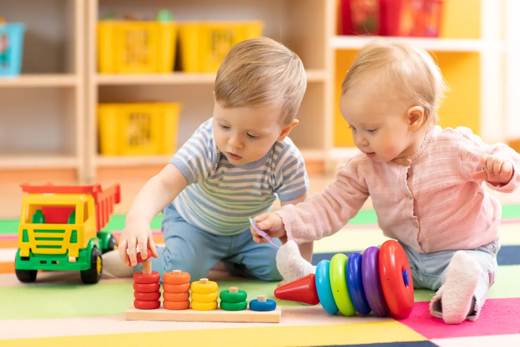 Zwei Kleinkinder spielen auf einer gemusterten Matte neben einem Spielzeug-Kipplaster mit bunten Stapelspielzeugen.
