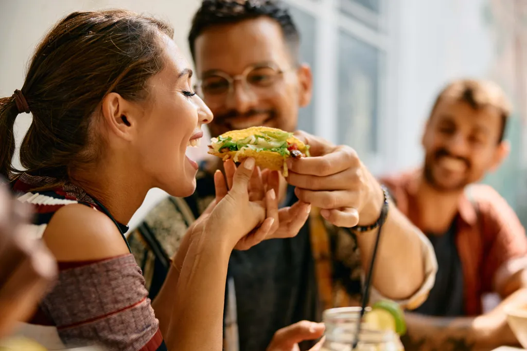 Eine lächelnde Frau, der ein Mann bei einem ungezwungenen Essen im Freien mit Freunden spielerisch einen Taco in den Mund schiebt.