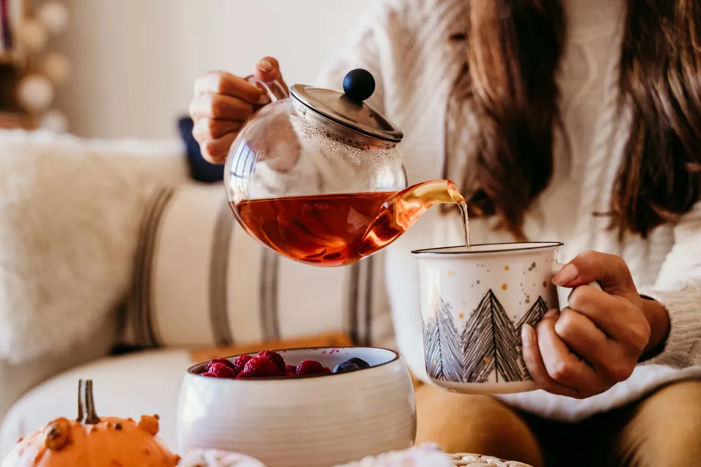 Eine Person, die Tee aus einer Glas-Teekanne in eine dekorative Tasse gießt, auf dem Tisch liegen Himbeeren und ein Kürbis.