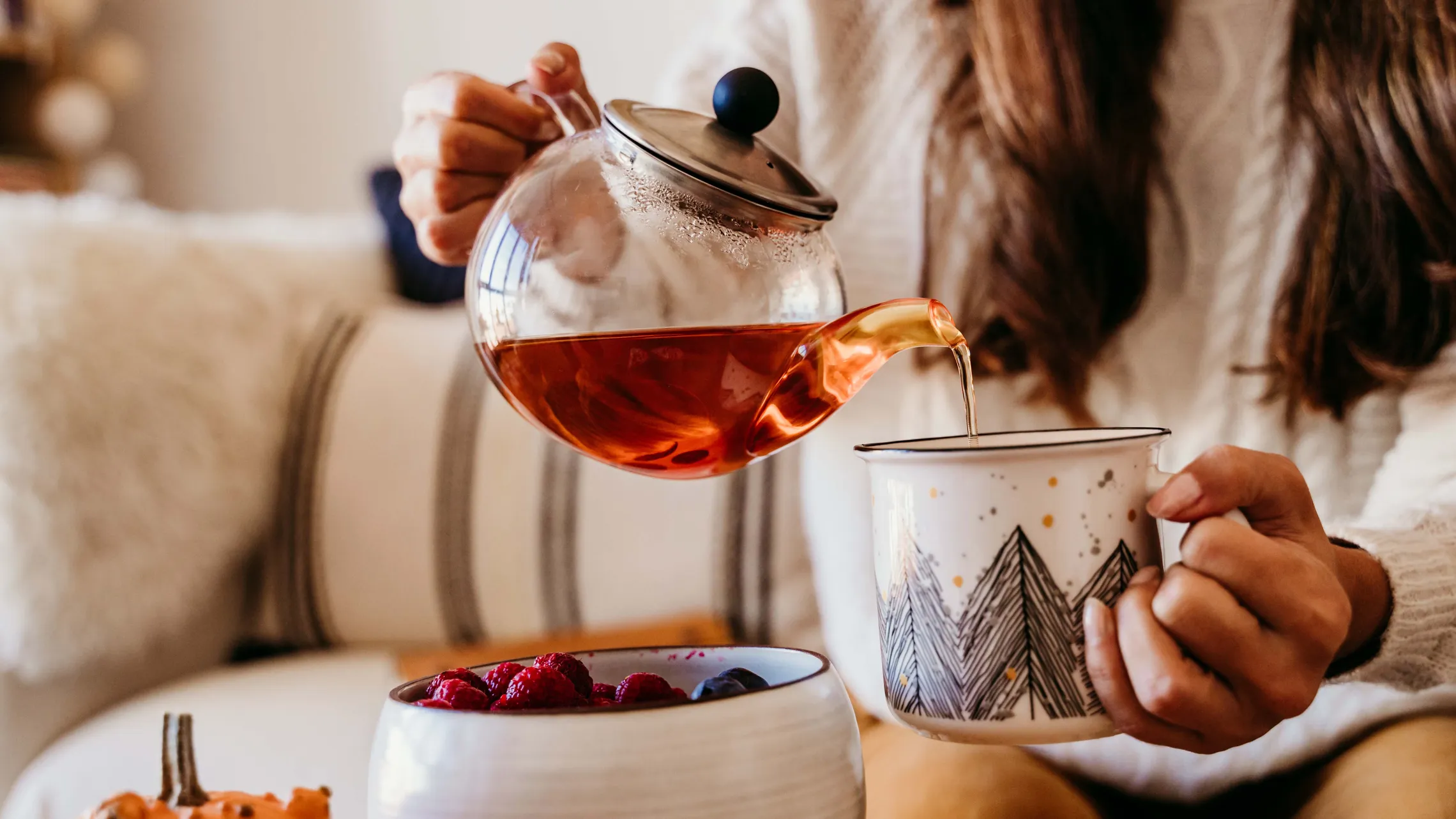 Eine Person, die Tee aus einer Glas-Teekanne in eine dekorative Tasse gießt, auf dem Tisch liegen Himbeeren und ein Kürbis.
