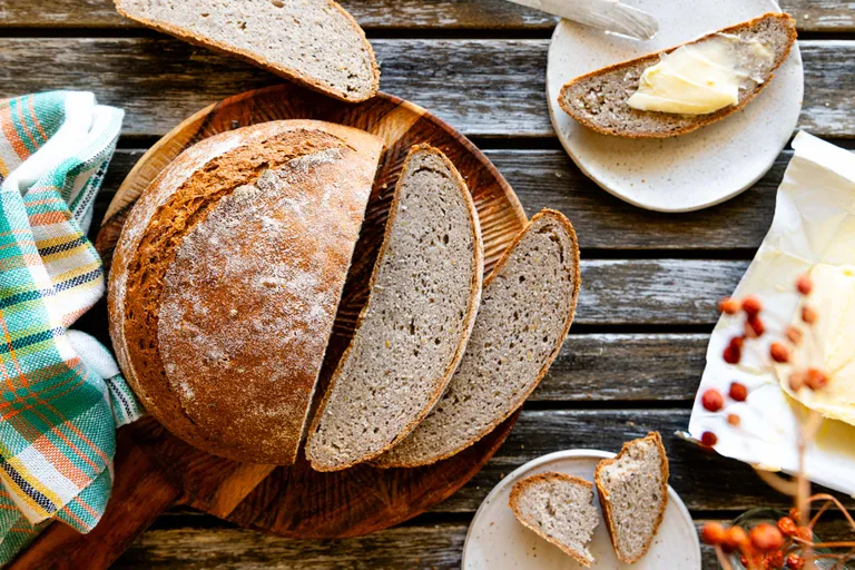 Ein aufgeschnittenes rustikales Brot auf einem Holzbrett, daneben auf einem Teller mit Butter bestrichene Scheiben, neben einer bunt karierten Tischdecke auf einem Holztisch.