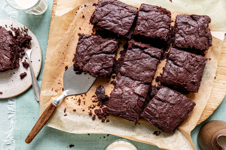 Neun vegane Schokoladenbrownies auf Backpapier, mit einem Spatel serviert, daneben ein Teller mit Krümeln und ein Glas Milch.