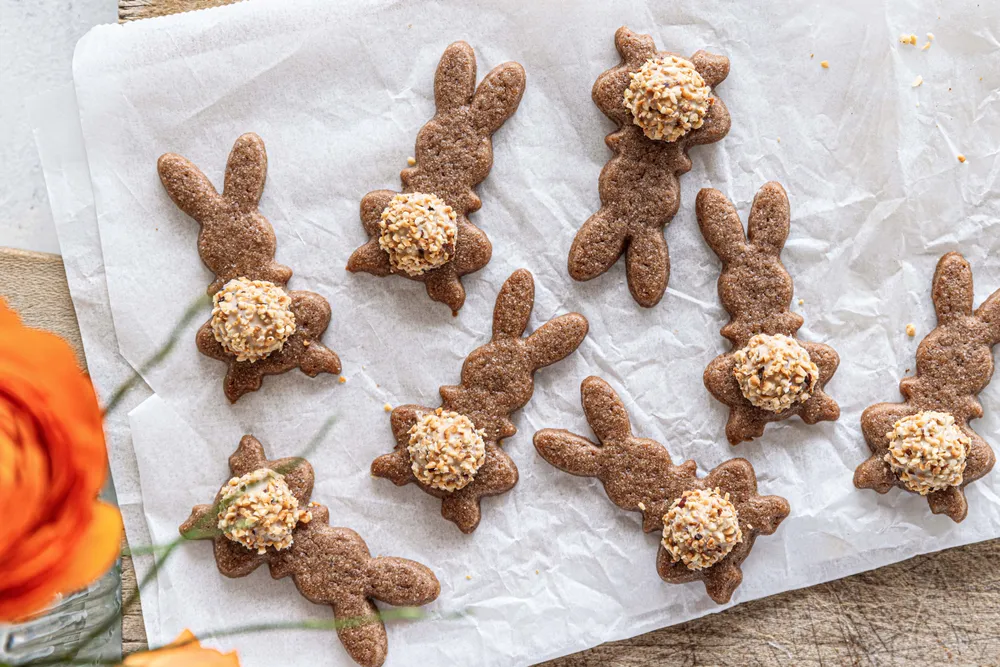 Hasen-Kekse mit Haselnuss-Schokoladen-Glasur auf Backpapier, umgeben von orangen Blüten.